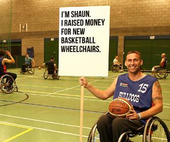 Shaun, in a wheelchair, holds a sign saying he raised money for basketball wheelchairs, with players in the background.