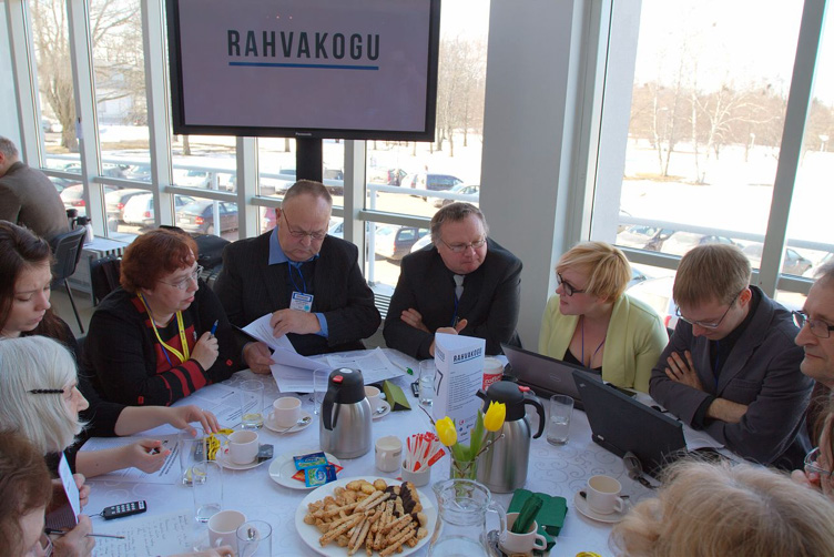 Group of people engaged in a meeting or workshop around a table, with a screen displaying "RAHVAKOGU" in the background.