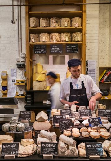 A cheesemonger in a Neal's Yard Dairy apron arranging various cheeses on a counter, with many labelled cheese types visible.