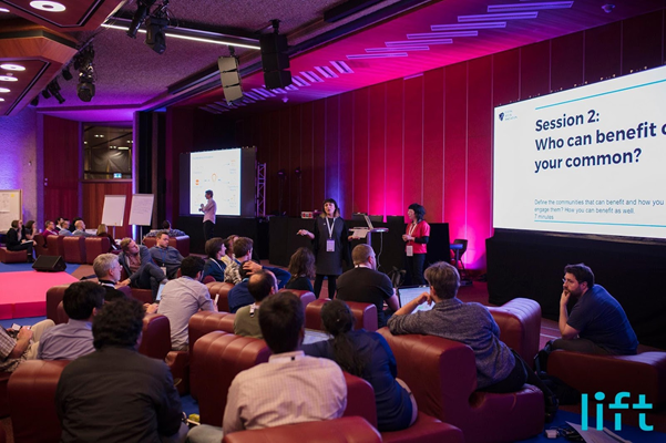 A conference session in progress, with speakers on stage and an audience, displaying "Session 2: Who can benefit your common?" on the screen.