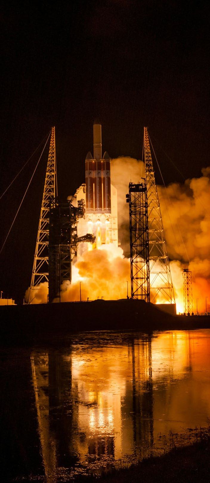 A rocket launching at night with bright orange exhaust flames and smoke reflected in a body of water.