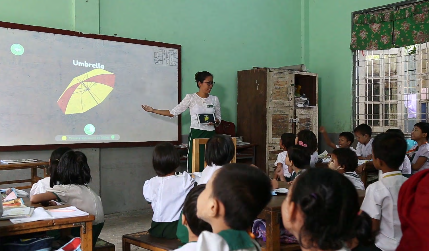 A teacher points to a projected image of an umbrella on a whiteboard, teaching young students in a classroom.