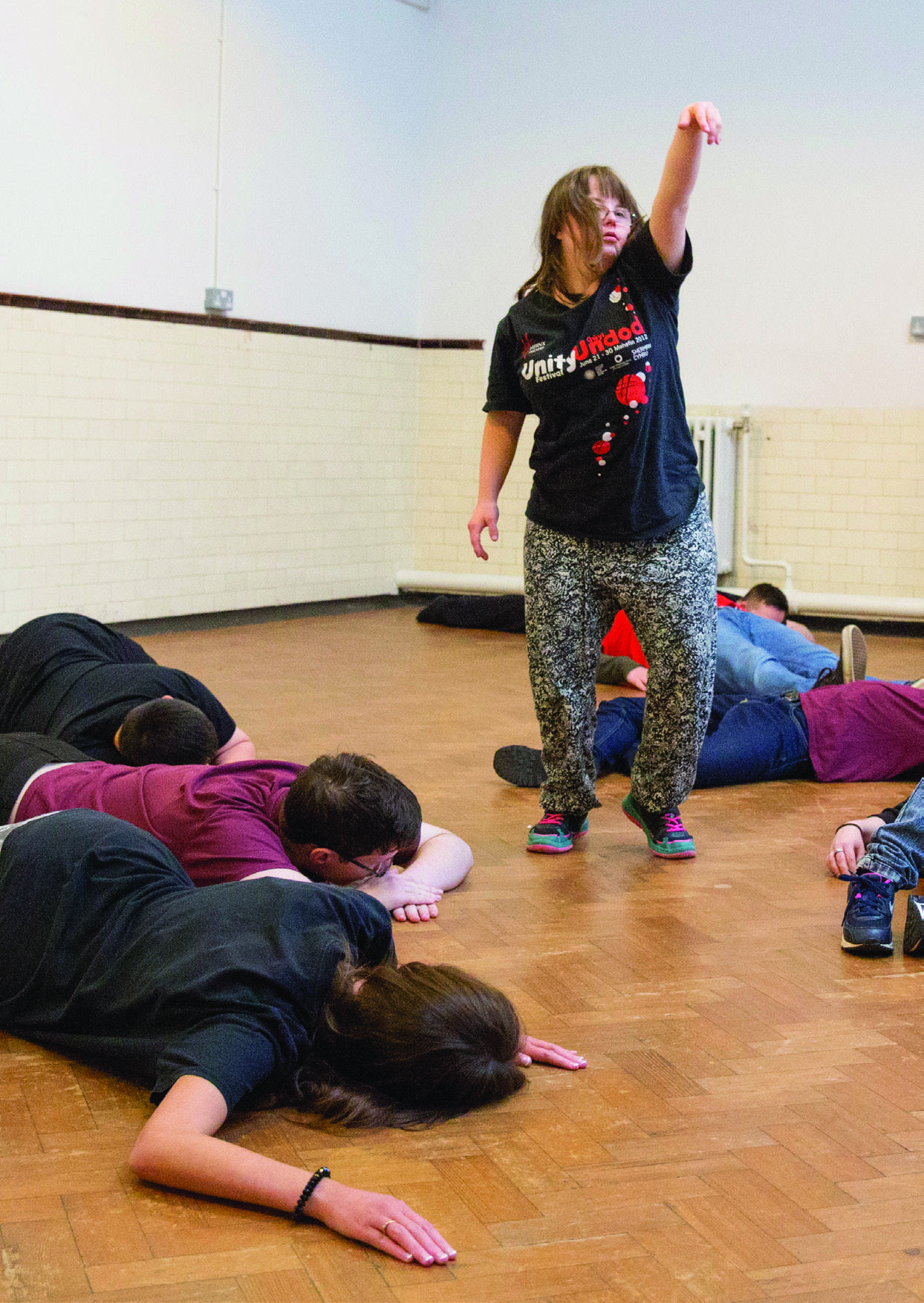 A person stands with arm raised while others lie on a wooden floor, appearing to participate in a group activity.