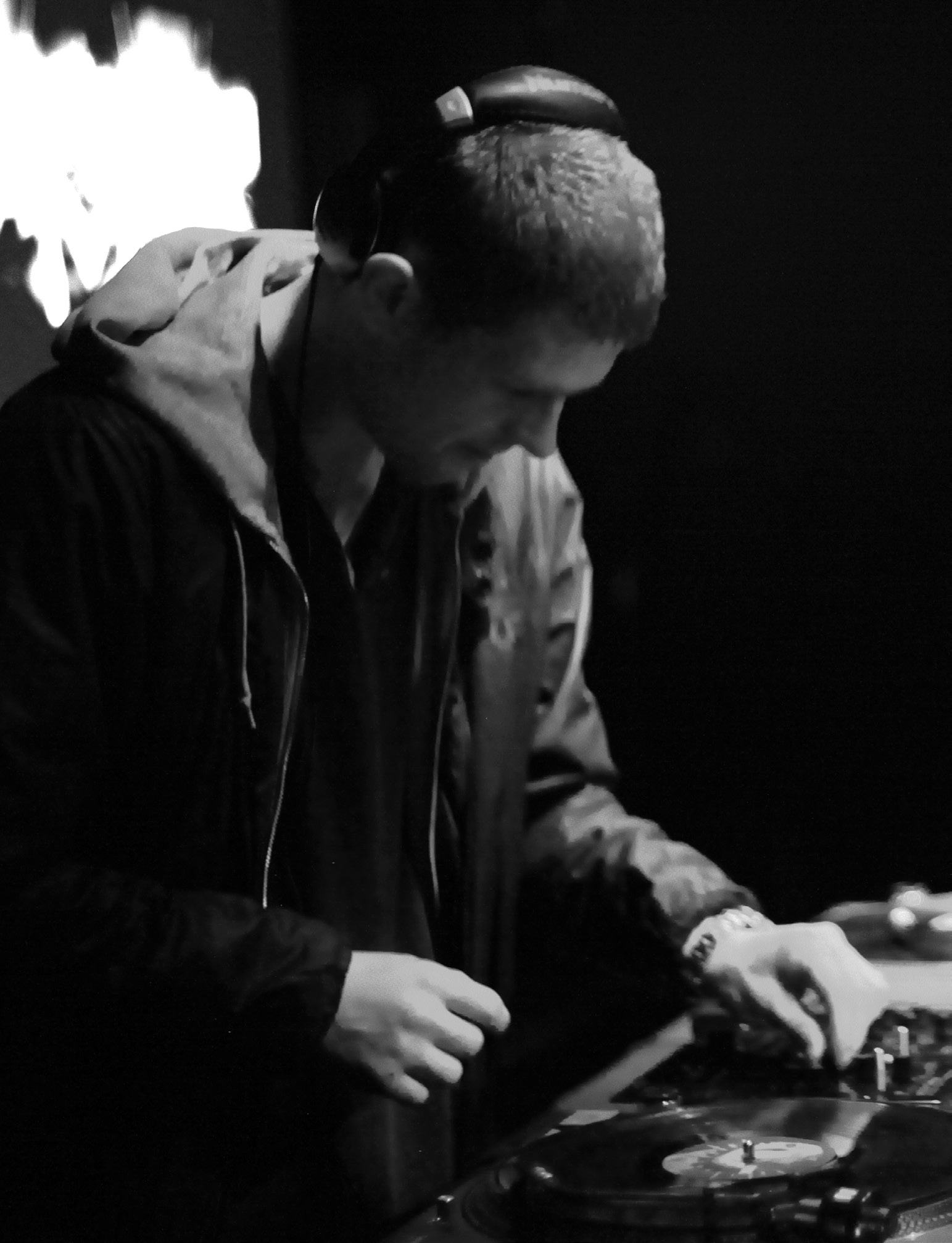 Black and white photo of a person wearing headphones, adjusting controls on a DJ mixer with a vinyl record.