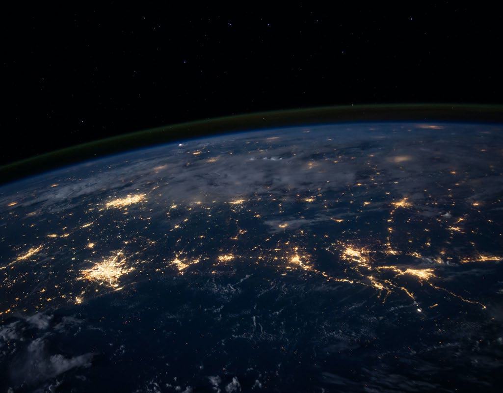 Earth seen from space at night, showing widespread clusters of city lights illuminating continents, with the dark sky above.