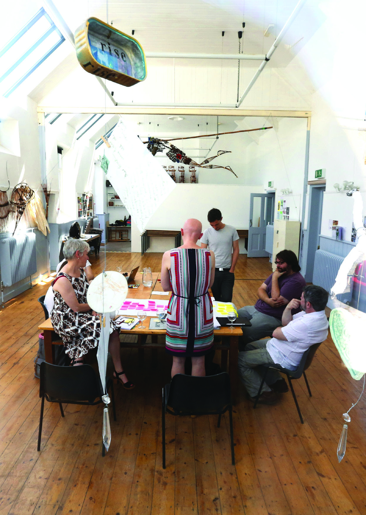 Group of people in a brightly lit room discussing around a table, with art installations hanging from the ceiling.