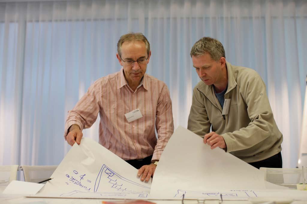Two men examining and unfolding large sheets of paper containing diagrams or architectural drawings during a meeting.