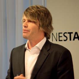 Close-up portrait of a man with shaggy blonde hair, wearing a suit jacket and white shirt, speaking at an event with Nesta branding.