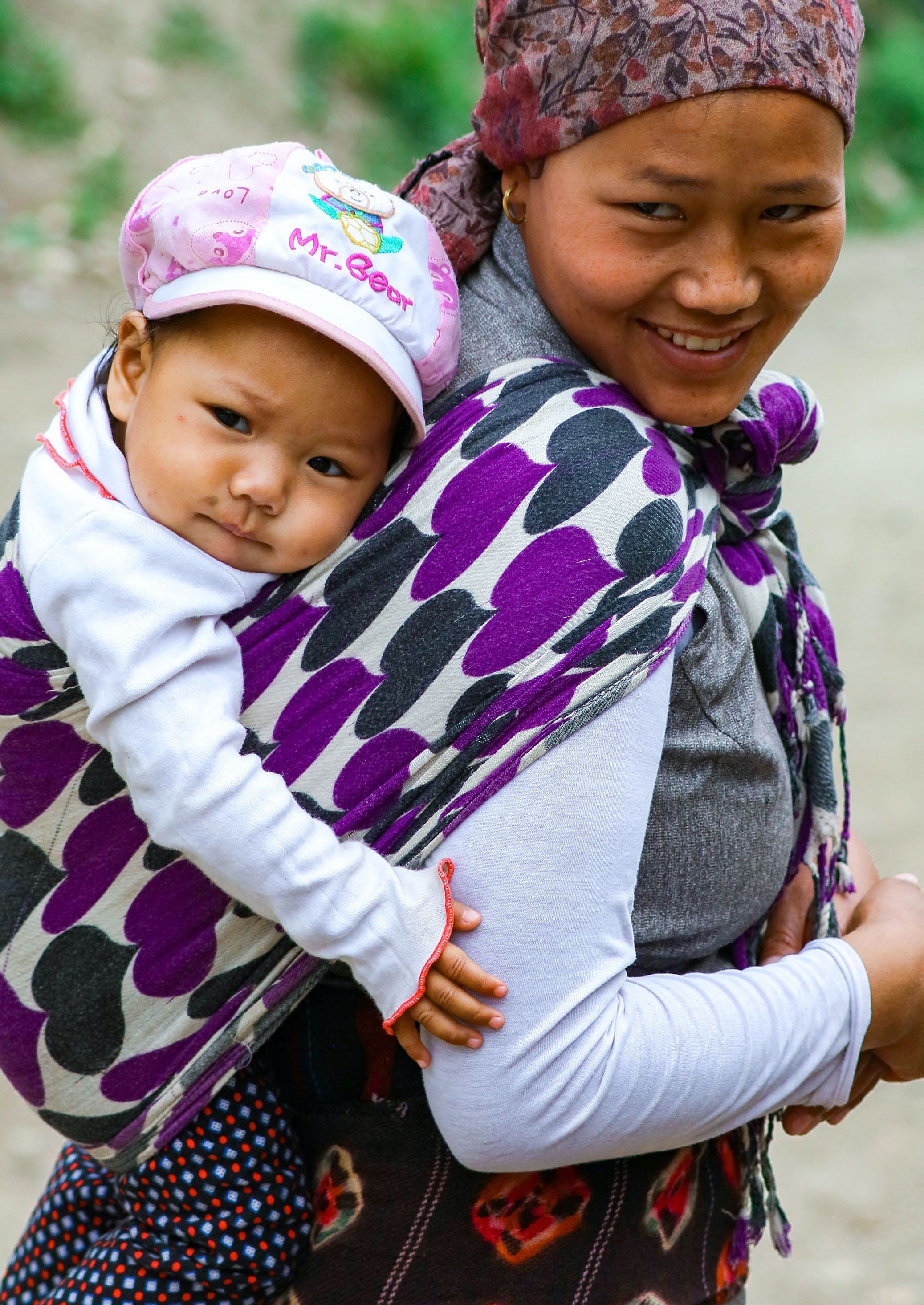 Close-up of a woman carrying a baby on her back in a patterned sling, both smiling.