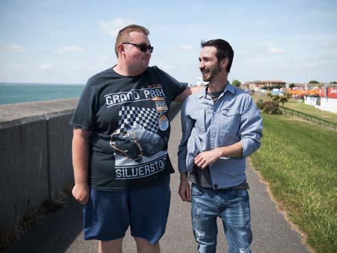 Two men walking on a paved path next to a sea wall, one with his arm around the other, conversing.