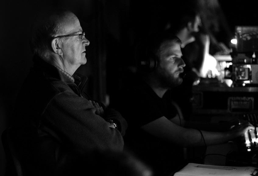 An older man and a younger man with headphones operating a control panel in a dimly lit technical setting.