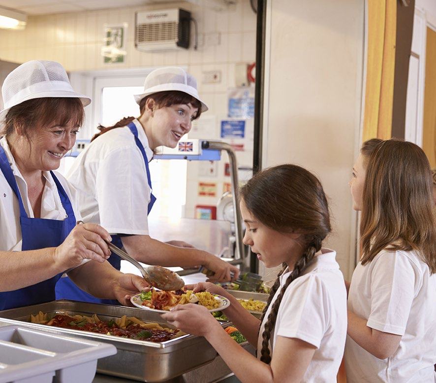 Two cafeteria workers wearing white hats and blue aprons serve food to two female students from buffet trays in a bright school cafeteria.