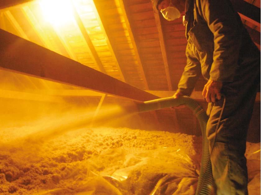 Person wearing a mask in an attic blowing insulation material with a hose between wooden beams.