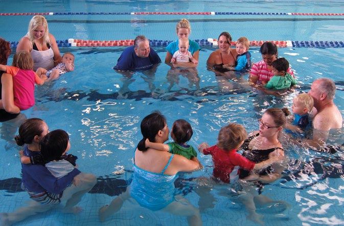 Group of adults and young children participating in a swimming class or therapy session in a pool.