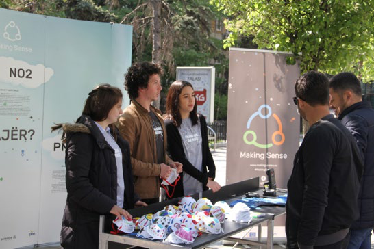 People at an outdoor 'Making Sense' project booth, discussing NO2 air quality with colorful masks on display, during a public engagement event.