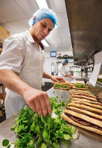 A chef in a hairnet adding greens to sandwiches in a commercial kitchen, with another chef working in the background.