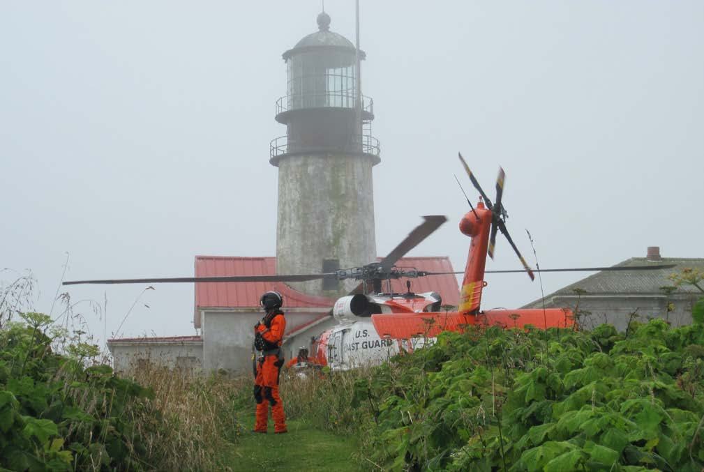 US Coast Guard helicopter and personnel in orange uniform near a lighthouse in heavy fog amidst green foliage.