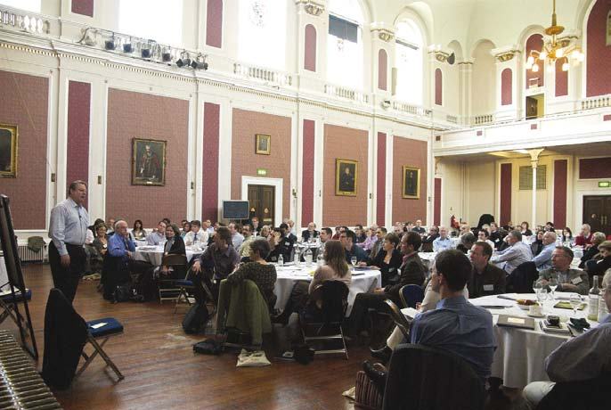 A man presents to a large audience seated at round tables in a conference hall.