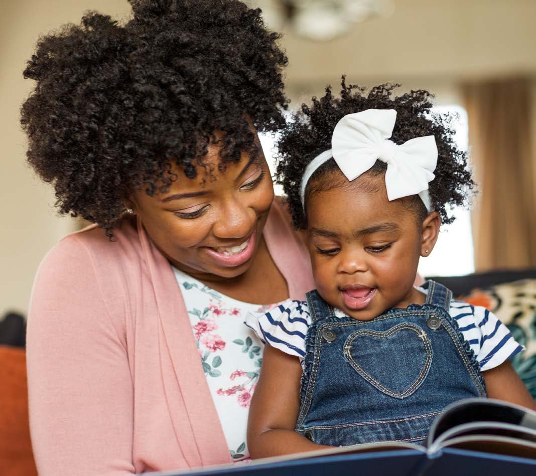 A woman smiling while reading a book to a young child with curly hair and a white bow, illustrating shared reading time.