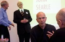 Attendees engaging in conversation and laughter at an event, with text 'JOHN SEARLE' visible in the background.
