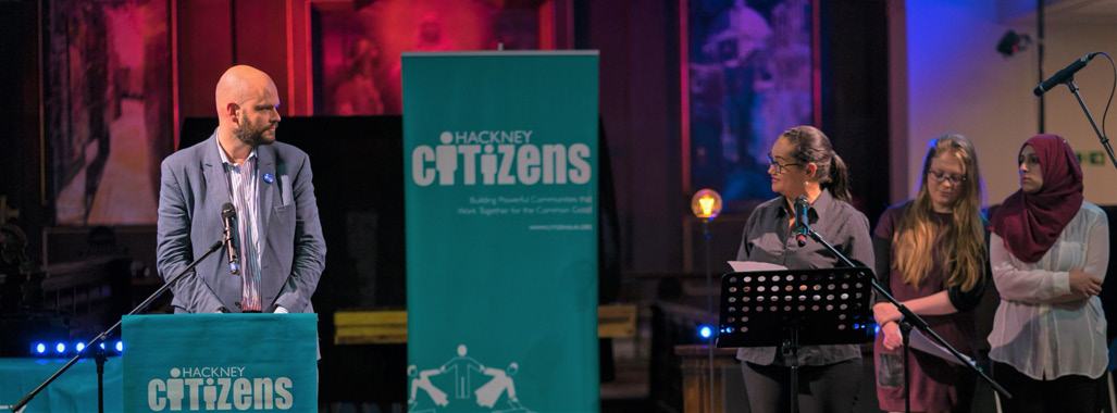 Man speaking at a podium with a 'Hackney Citizens' banner, three women standing nearby on a stage, suggesting a community event.