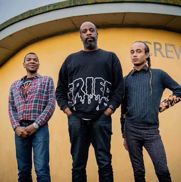 Three men of varying ages pose in front of a yellow curved building with a sign that reads "CREW".