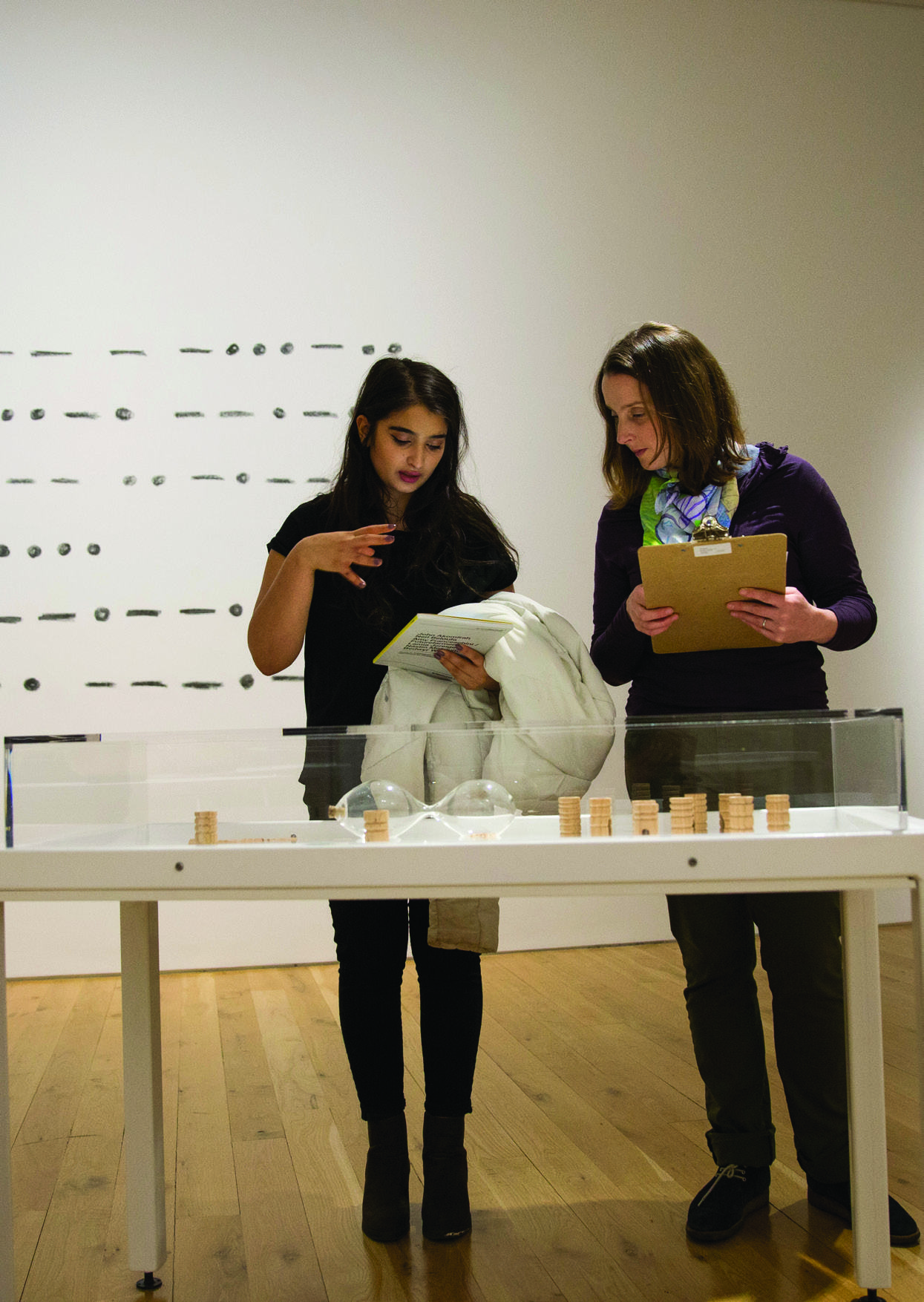 Two women observing objects in a clear display box on a table, with abstract patterns on a white wall in the background, likely at an exhibition.