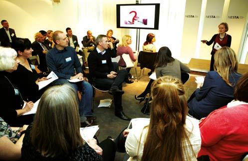 An audience attending a presentation with a speaker at the front and a screen displaying a red question mark graphic.