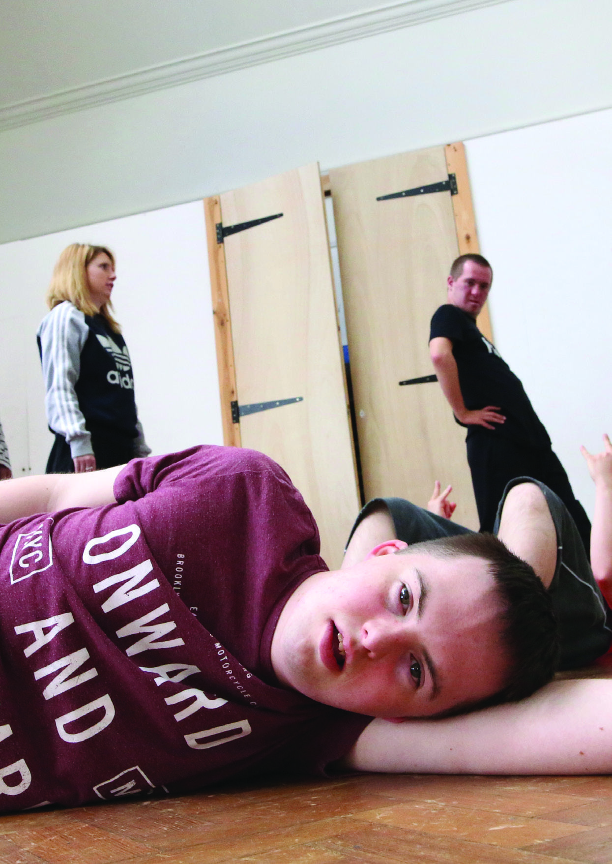 A young person with Down syndrome lies on a wooden floor, looking up, with two adults standing in the background.