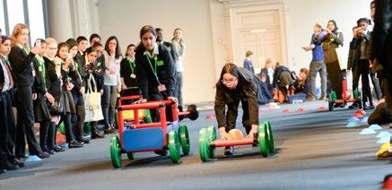 Students in uniform observe two peers racing small, human-powered vehicles in a large hall, part of an educational event.
