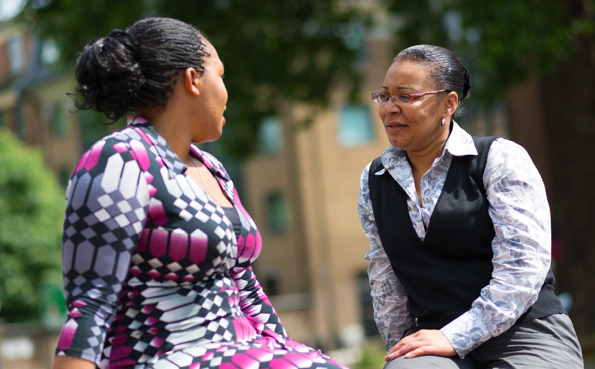 Two women in conversation outdoors. One wears a pink and black patterned top, the other a white collared shirt with a dark vest and glasses.