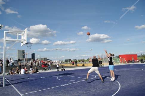 People playing basketball on a blue outdoor court in a city park under a bright blue sky with clouds.