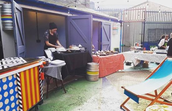 Outdoor food market scene with several stalls, a vendor, and customers in an open area.