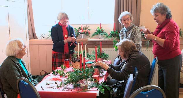 Group of older women sitting and standing around a table, engaged in decorating with candles and greenery.