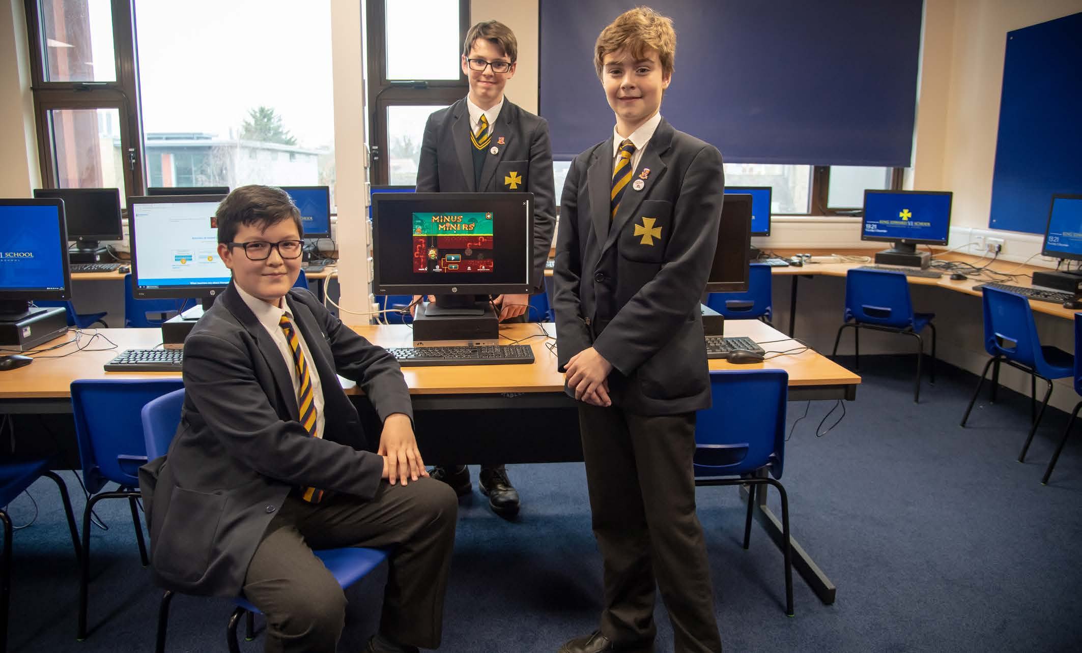 Three male students in a computer lab, with one computer displaying the "Minus Miners" game, all looking at the camera.
