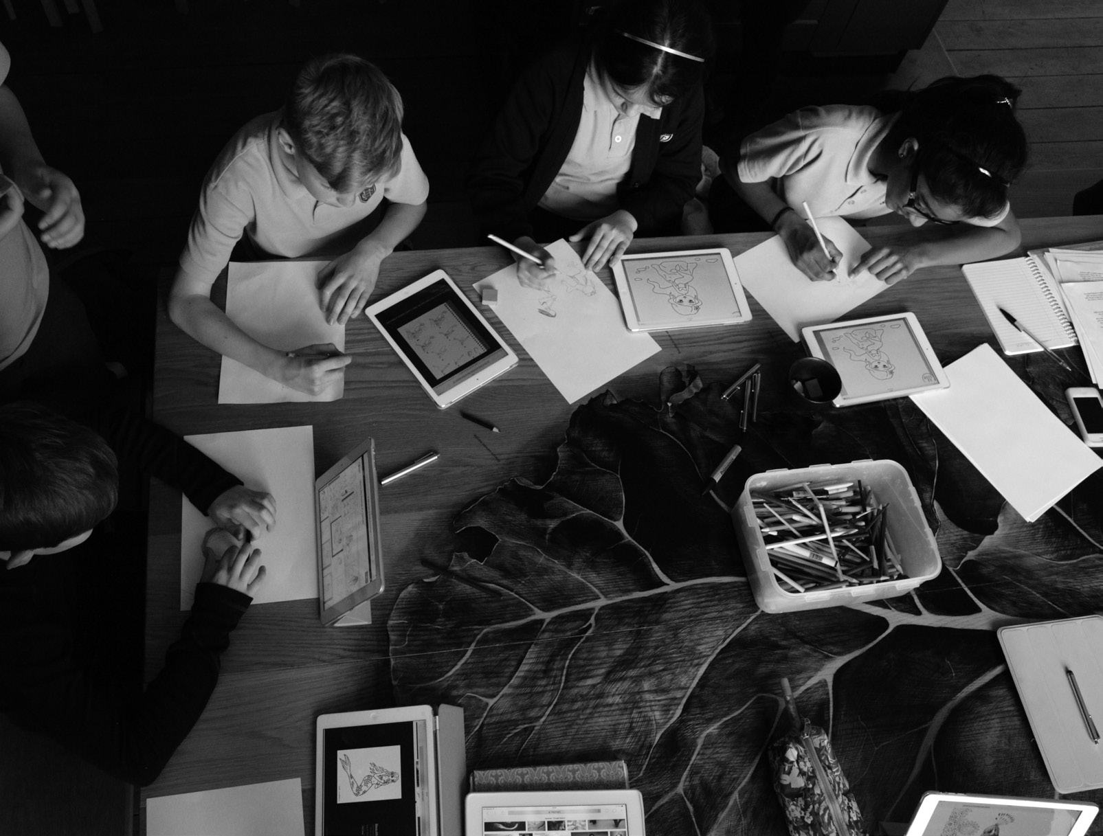 Overhead view of students working collaboratively at a large table with papers, art supplies, and digital tablets.