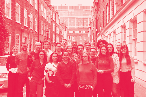 A large group of diverse people, likely a team, stand together on a street in front of brick buildings.