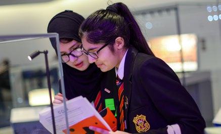 Two students in school uniforms, one wearing a hijab, examining a document under a spotlight in an educational setting.