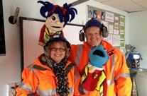 Two smiling people in orange high-visibility gear and hard hats, posing with two puppets, possibly from a safety or educational program.