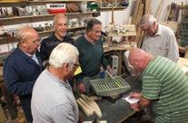Group of senior men gathered around a workbench in a workshop, observing one man taking notes on a clipboard.
